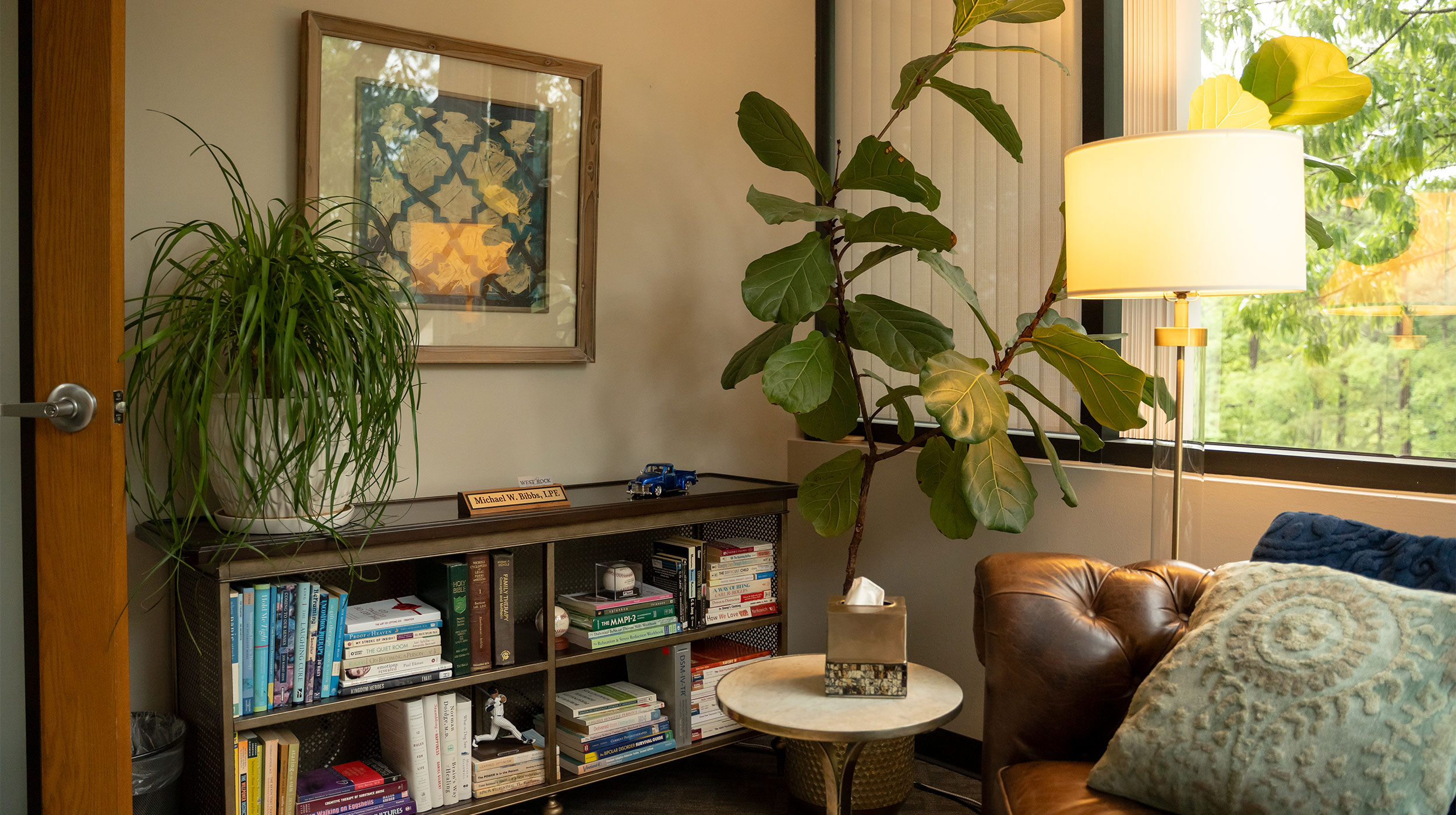 WRC-Background A cozy office corner with a leather sofa, a patterned pillow, a small round table with a tissue box, a large leafy plant, a bookshelf filled with books, and a lamp by a window letting in natural light.