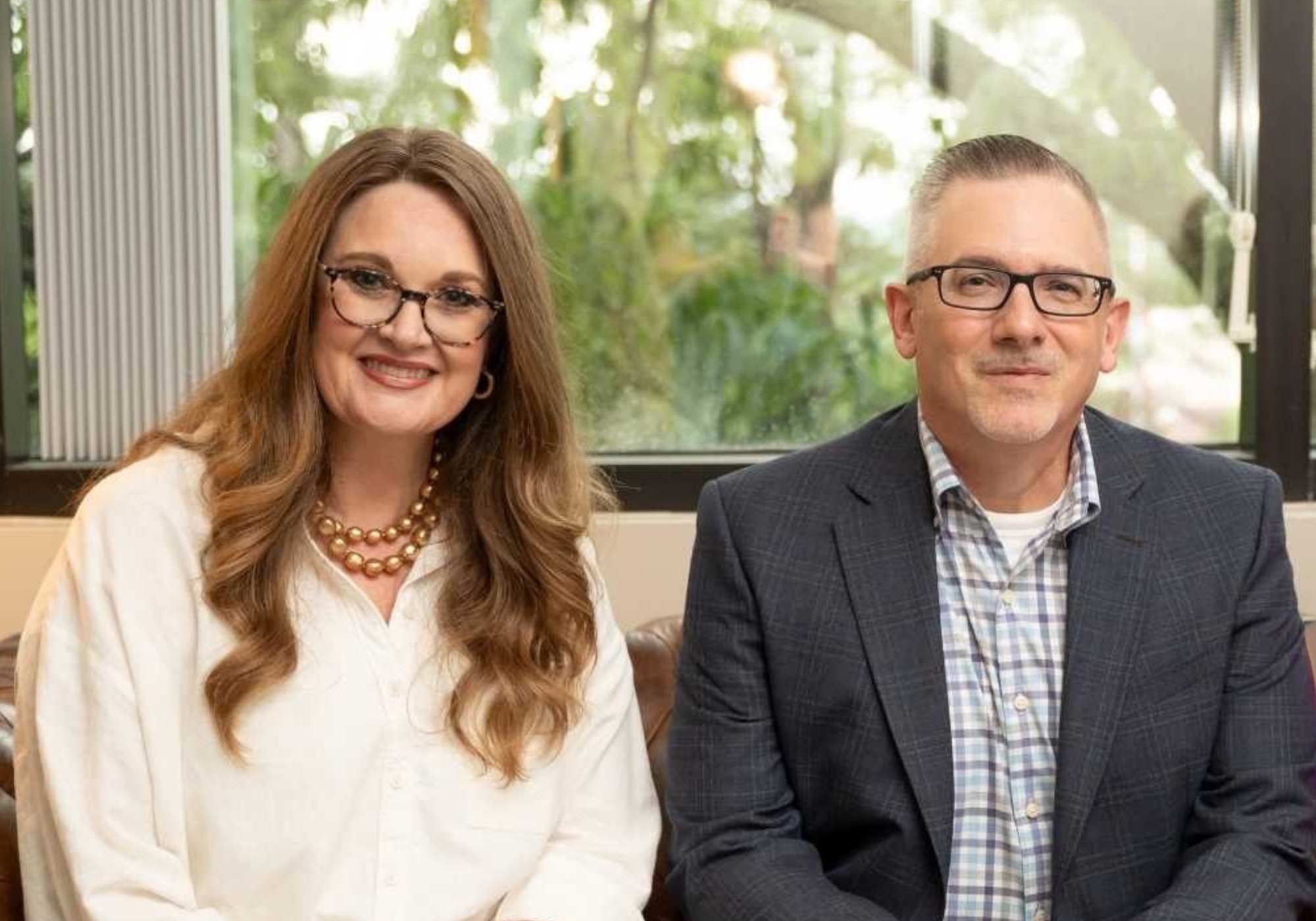 A woman with long wavy hair and glasses sits next to a man in a plaid shirt and blazer, both smiling, in front of a large window with greenery outside.