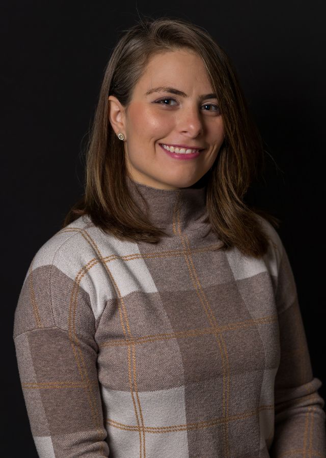 Tara Hamilton A woman with straight brown hair and a plaid turtleneck sweater smiles at the camera against a plain black background.